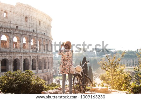 Beautiful young woman in colorful fashion dress alone on a hill with bike takes pictures of colosseum in Rome using smartphone camera at sunset. Attractive tourist girl with elegant straw hat.