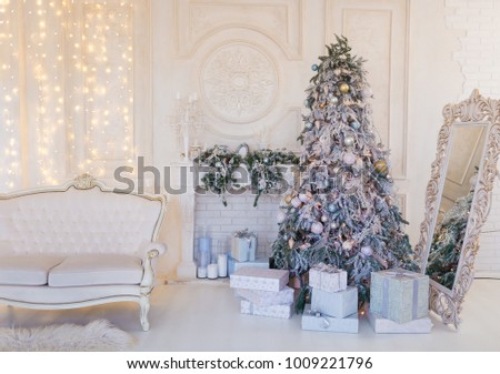 Silver Xmas tree  near sofa and gift boxes near it in white room with fireplace, wall decorated by lights 