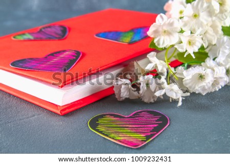 Black and pink hearts on a book in a red cover and flowers on a gray concrete background. The concept of Valentine's Day. A symbol of love