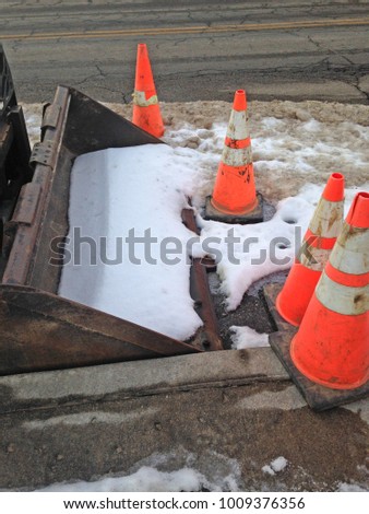 An excavation equipment piece has snow in the bucket and is surrounded by orange construction cones.