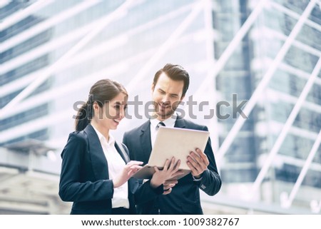 Two young contemporary businessmen and Asian businesswomen working outdoor using a smart phone and a notebook laptop with new startup project , both smiling - technology, business, working concept
