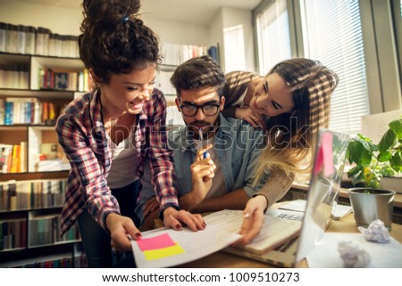 Close up view of two adorable playful attractive hardworking high school student girls hugging their confused handsome nerd bearded classmate and asking for fun in the sunny library. Royalty-Free Stock Photo #1009510273