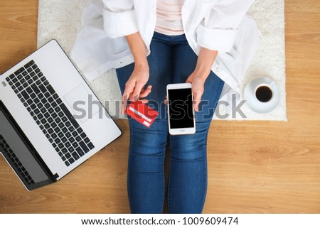 Top view of young woman with laptop & cellphone on her lap, sitting on wooden floor background. Overhead, female hands on keyboard, holding credit card, notebook computer, mobile gadget, coffee cup.