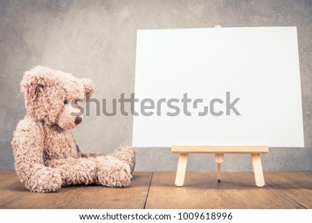 Sitting old Teddy Bear toy near portable desk easel for painting with canvas blank front concrete wall background. Retro old style filtered photo