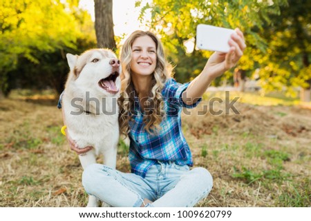 young stylish pretty smiling blond woman playing with dog in park, happy, husky breed, summer style, sunny, positive mood, making selfie photo on phone