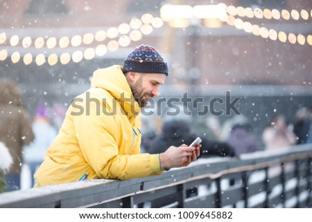 Portrait of joyful bearded man in yellow jacket, black hat standing near fence on ice rink and using smart phone, outdoors at snowy day/ Winter time concept/ Weekend activities outdoor in cold weather