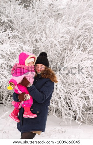 Adorable laughing baby girl in a warm pink snow suit playing in snow on a winter day in a forest with her mom