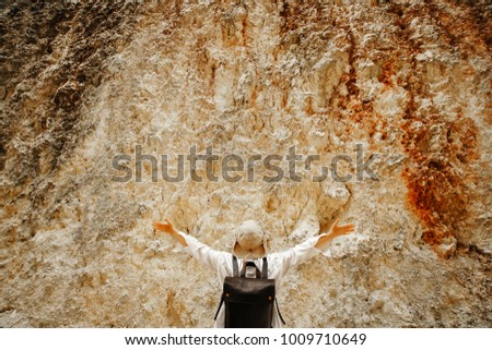 girl with a white shirt and a leather backpack is standing on a stone wall with her hands up. Delight.