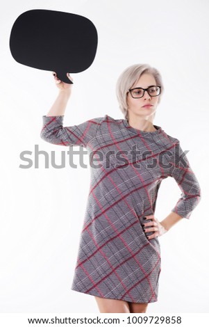 Young beautiful emotional blonde woman in black glasses is thinking about some things and holding speech bubble cutout on white background. Three quarters body photo