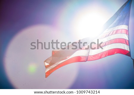 American Flag flying to the Left with Solar Flares and lens flares shining right into the camera and bright illuminated Large American Flag high up in the blue sky