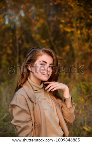 Stylish and beautiful red haired girl posing, walking in autumn forest among yellow grass. Woman with long hair, in jeans, blouse and brown coat, smiling, relaxing outdoors.