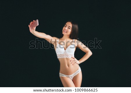 Portrait of a smiling cute woman making selfie photo on smartphone isolated on black background. Young woman with long black hair stands isolated on black background