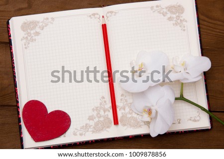 open blank notepad for notes with red pencil and red heart beside white orchid flowers on a wooden table
