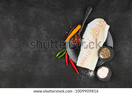 Cod fillets on a black ceramic plate for the preparation of a healthy dish with the addition of pink pepper, hot pepper, spinach, lemon, salt on a simple black background. Top view. Copy space