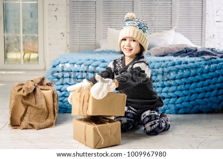little girl a child in hat and mittens folds a mountain from the boxes of gifts at home,near the bed.The interior is decorated with Christmas decor.Daytime bright sunlight from window. New Year theme