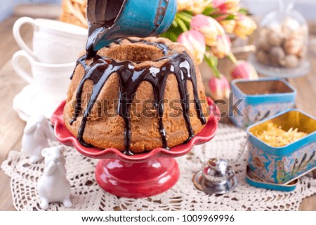 Homemade sweet cake, poured with chocolate, on a red cake plate. On a brown wooden background, dry herbs for tea and a cup. A bouquet of spring flowers. Tulips and a ceramic rabbit. Postcard and free 