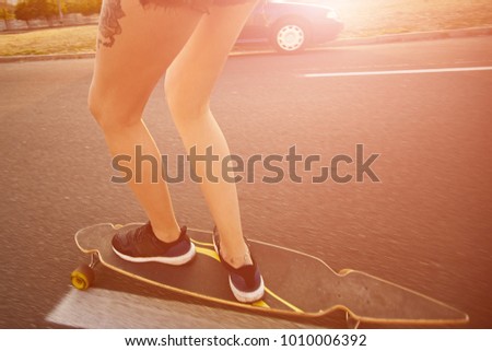 Girl legs rolling a longboard on the city road in the sunlight with urban life in background. Cropped image of hipster with tattoos skateboarding in the city.