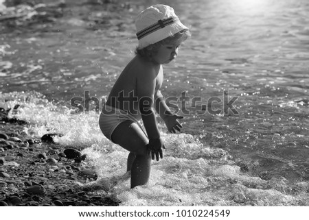 Little curious funny blonde baby boy in hat sitting on sand sunny day outdoor playing with wavy ocean water on natural beach background, horizontal picture