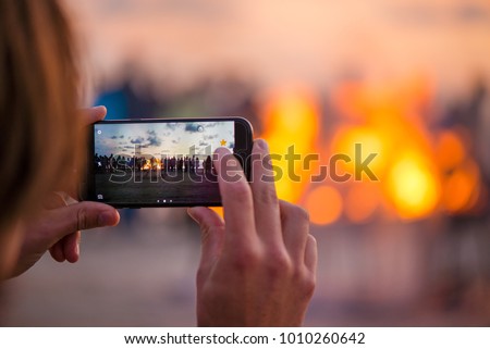 Woman is taking picture of sunset with smart phone. Romantic bonfire night at seaside. People gathering together to celebrate Night of ancient lights. Large burning campfire with soft glowing flame.