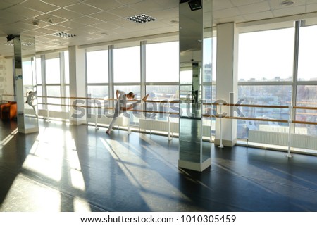 Gymnast train near ballet barre in sport gym, guy diligently doing stretching exercises for legs. Tattooed concentrated male wearing white sportswear sloping. Concept of well-equipped hall, sport