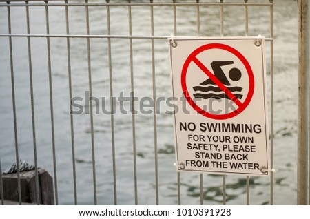 Warning sign for "No swimming for your own safety please stand back from water" at the fence near waterfront.