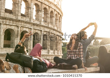 Three young friends tourists sitting lying in front of colosseum in rome taking selfie pictures with smartphone camera. Sunset with lens flare.