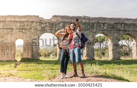 Young couple backpackers tourists with guitar taking selfies in front of roman aqueduct ruins in acquedotti park in rome.