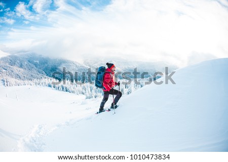 A man in snowshoes and trekking sticks in the mountains. Winter trip. Climbing of a climber against a beautiful sky with clouds. Active lifestyle. Climbing the mountain through the snow. Royalty-Free Stock Photo #1010473834