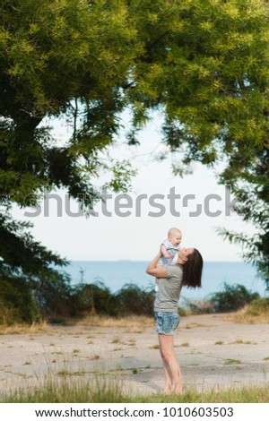 Young fun smiling woman standing at sea background hugging little cute child baby boy on nature, green trees. Mother, little kid son. Parenthood, family day 15 of may, love, parents, children concept