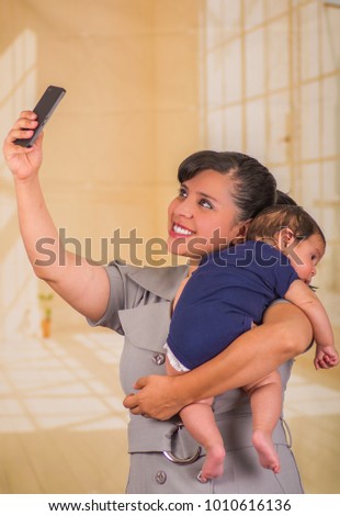 Young mother is holding her little baby and taking a selfie, in a blurred background