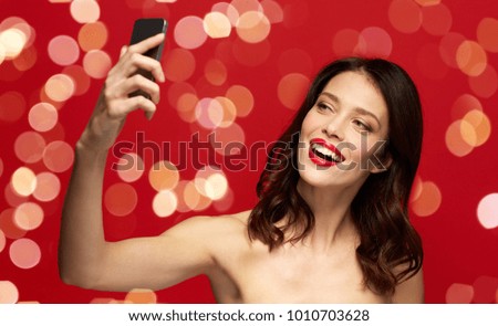 beauty, make up and people concept - happy smiling young woman with red lipstick taking selfie by smartphone over red background with lights