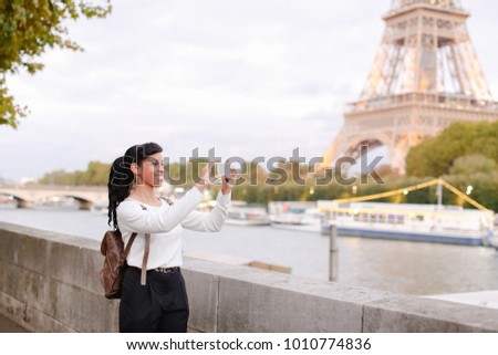 Student walking along embankment not far from Eiffel Tower and making photos of landmarks using smartphone. Young woman recently moved to Paris to live and study. Smiling black-haired female with
