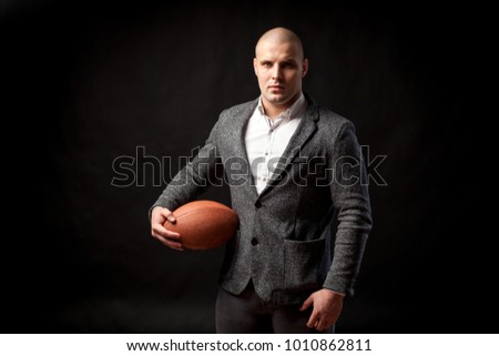 A young bald man in a white shirt, gray suit holds a rugby ball in one hand and poses on a black isolated background