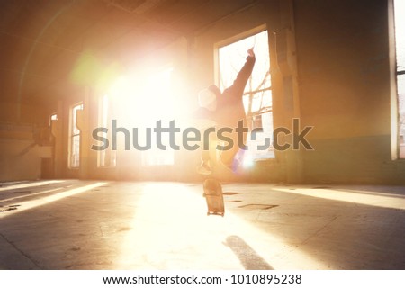 A young skater in a white hat and a black sweatshirt does a trick with a skate jump in an abandoned building in the backlight of the setting sun.