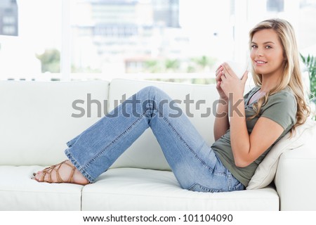 A woman sitting sideways on the couch, legs raised slightly, smiling and a cup raised to her mouth.