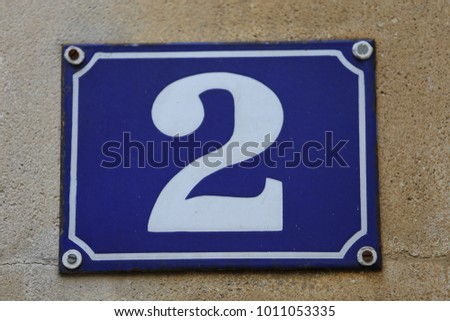 Number two on a metal plate. 2 written in white in a blue rectangle with a white frame. Sign of the adress of an house. Plate placed on a beige stone wall near a wooden door. Abstract urban picture  