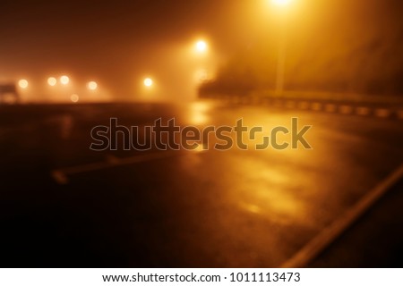 Blurred background of night time empty carpark shrouded in fog with warm street light.