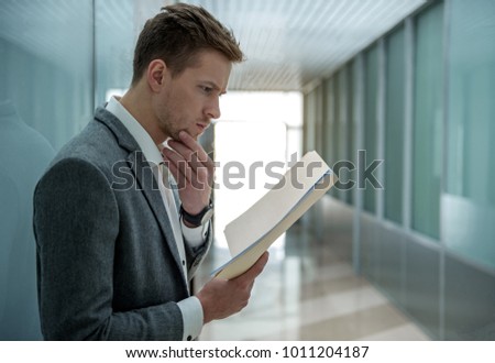 Busy morning. Pensive young professional employee is reading documents with concentration. He is standing in hallway and touching his chin. Copy space in the right side