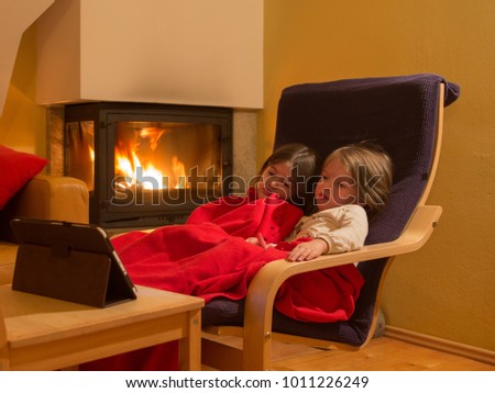 two little sisters sit by the fireplace on a winter evening and watch cartoons on a tablet