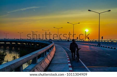 People relaxing on curved coastal road with street lamp and orange sky at sunset in Chonburi, Thailand.