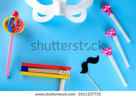 Happy Purim Jewish celebration holiday greeting card. White carnival mask, colorful wooden and paper noisemakers or graggers and cute mustache on blue background.Copy space for text.Top view.Flat lay.