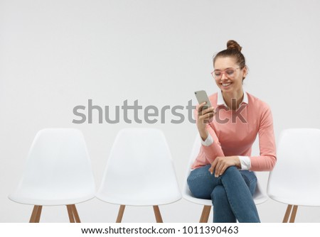 Woman wearing eyeglasses and casual pink sweater sitting in the line, holding smartphone texting sms. Busy woman sitting on chair indoors