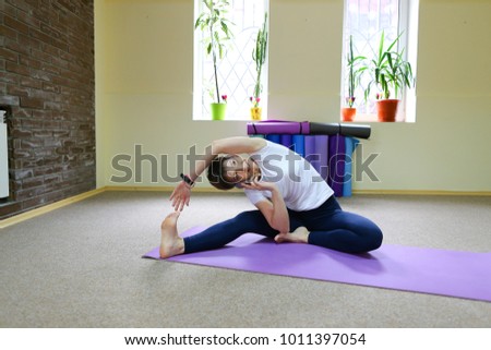 Beautiful young woman of American origin stands in middle of room on yoga mat and performs asanas standing on one leg. Girl dressed in sporty black tights and white T-shirt, hair is black and short