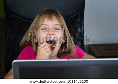 Little girl sitting at the computer and eating chips.