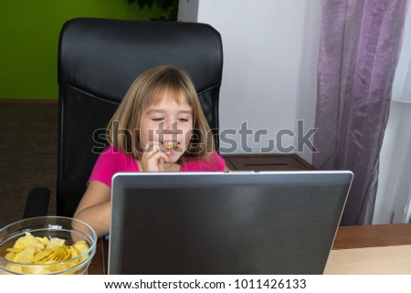Little girl sitting at the computer and eating chips.