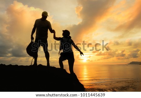 Male and female hikers climbing up mountain cliff looking at beautiful sunset  and one of them giving helping hand . People helping and, team work concept.
