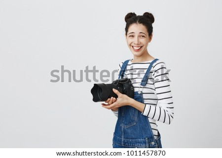 Smiling joyful positive european brunette girl photographer with two hairbuns wearing denim overall using modern camera. Trendy young female smiling posing with camera against studio background