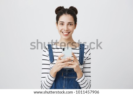 Happy brunette young woman with hair knots, wears denim overall, uses smartphone for taking pictures, smiles pleasantly, isolated against gray background. People and happiness concept