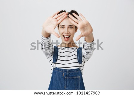 Cute joyful smiling young beautiful female in denim overalls stretches arms to camera, demonstrating her white teeth, having fun indoors, rejoicing positive news, isolated against gray background.