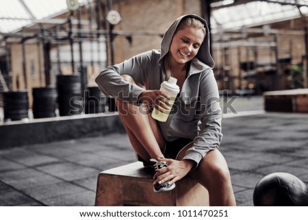 Sporty young blonde woman wearing a hoodie smiling and drinking water while sitting on a box at the gym after a workout 